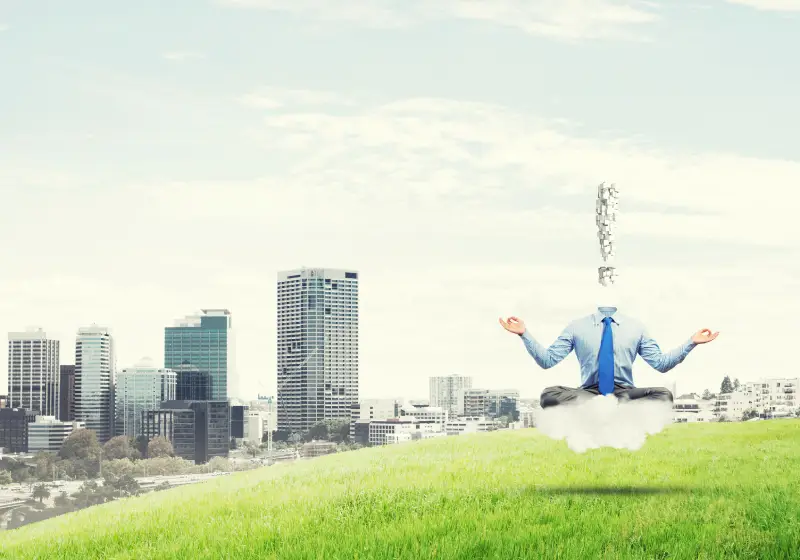 A photo of a headless man in a business shirt and tie, sitting in a meditative pose on a white cloud in a grassy field. An exclamation point made of interconnected wireframe lines is floating above his neck. A city skyline is visible in the background under a cloudy sky.
