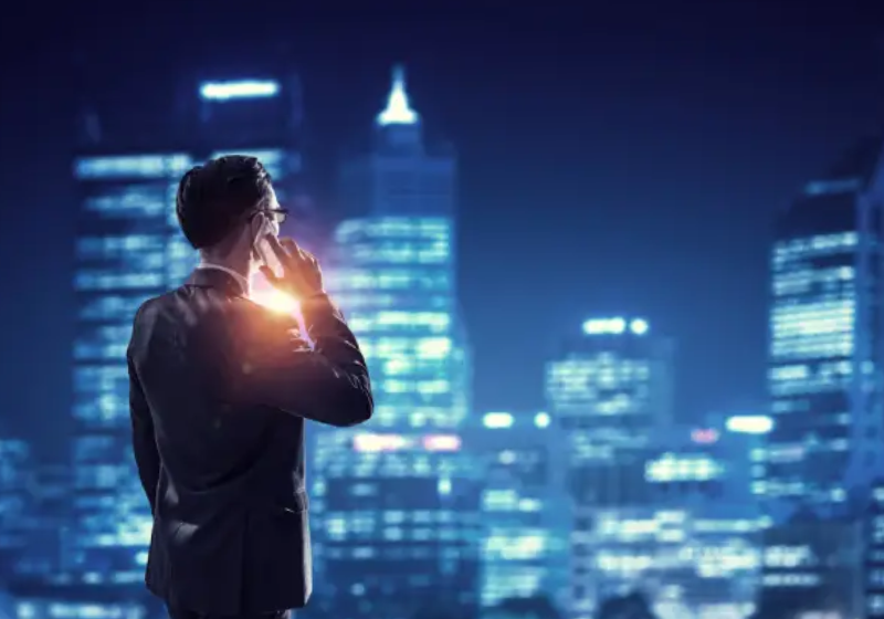 A low-angle photo from behind a businessman in a suit, looking out at a city skyline at night. He is holding a phone to his ear with his left hand, which is slightly illuminated. The city lights are blurred in the background, creating a glowing effect.